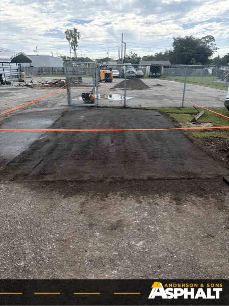 Freshly laid asphalt patch with compacted base material and skid steer visible beyond a chain-link fence