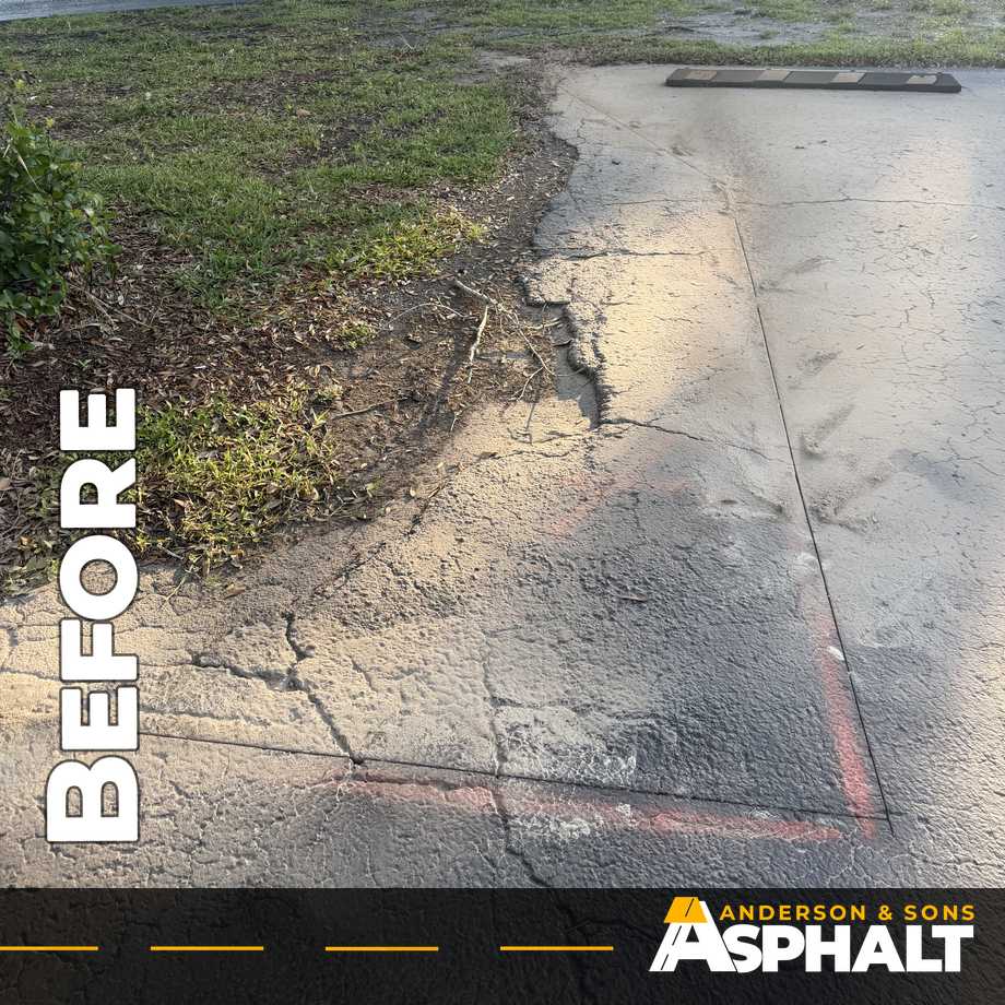 Cracked asphalt crumbling into the grass along a landscaped island, repair area marked in red paint